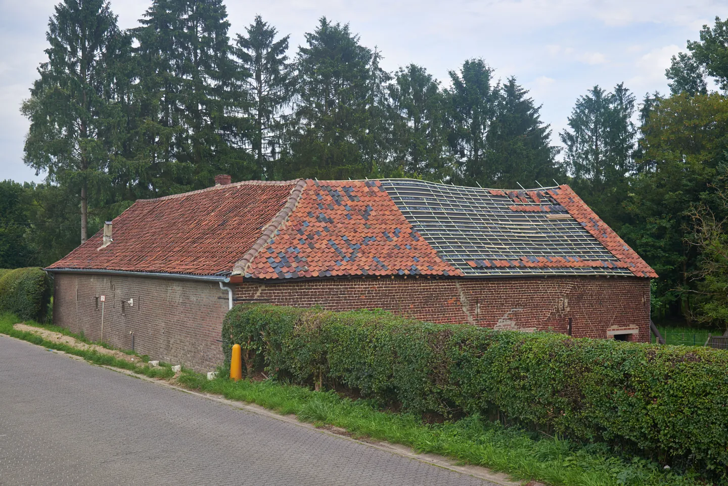 Alacsony téglaépület részben hiányzó cserépfedéssel, látható tetőlécekkel.