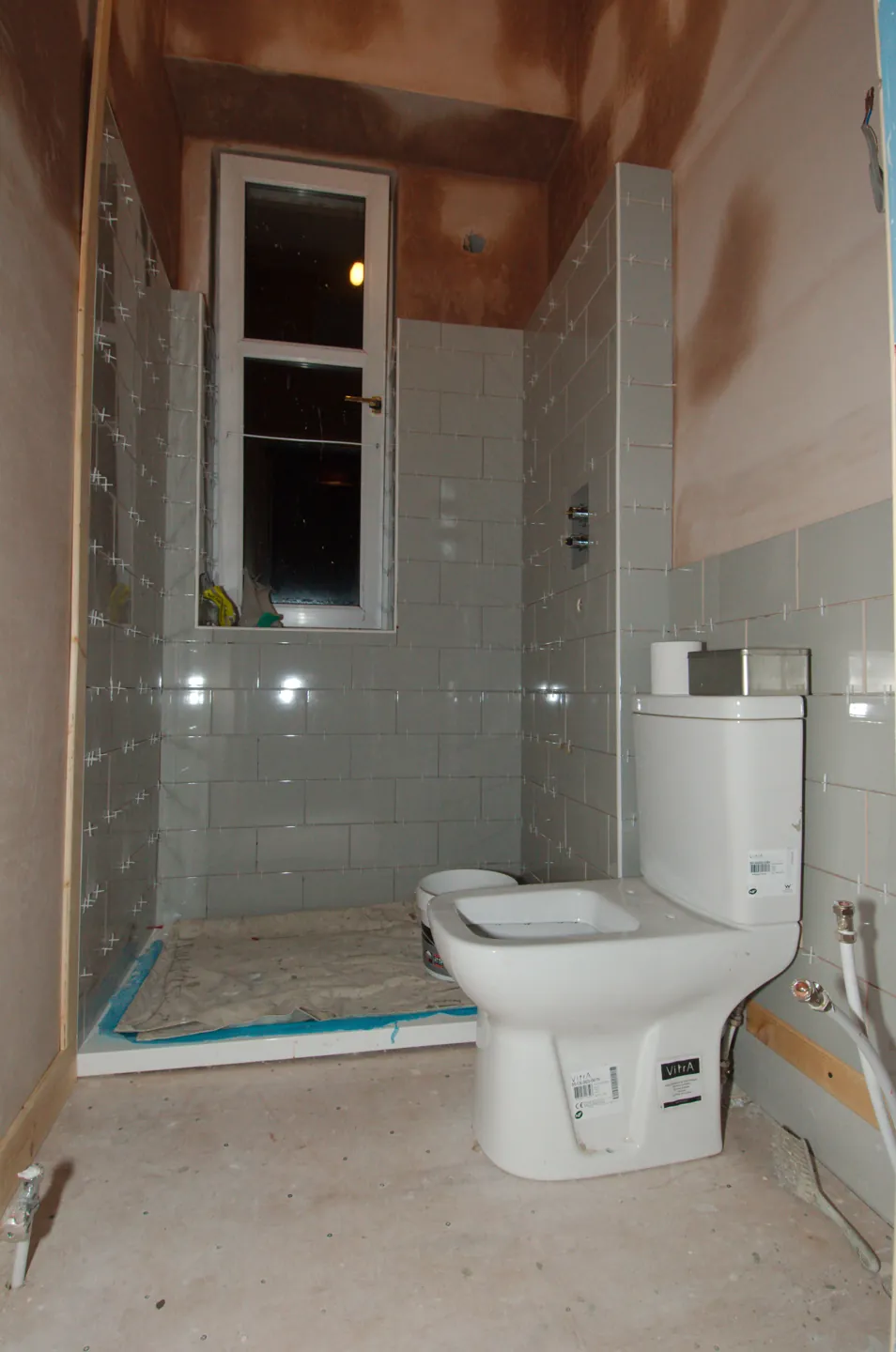 Részben burkolt fürdőszoba zuhanyaljjal, WC-vel és még nyers felső falszakaszokkal.
