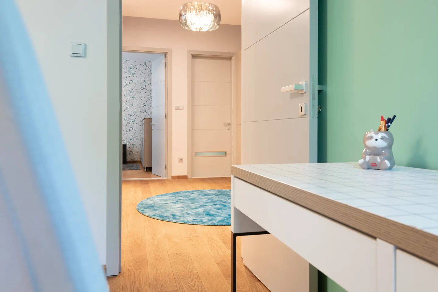 Nyitott világos beltéri ajtón át látható közlekedő; előtérben rácsmintás íróasztal, ceruzatartó és zöld fal.
