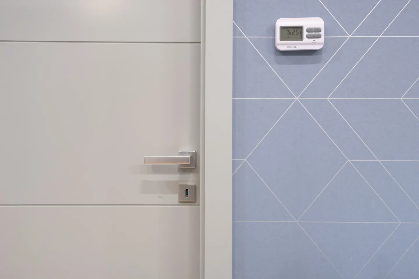 Fali digitális termosztát kék geometrikus falon, világos beltéri ajtó és fém kilincs mellett.