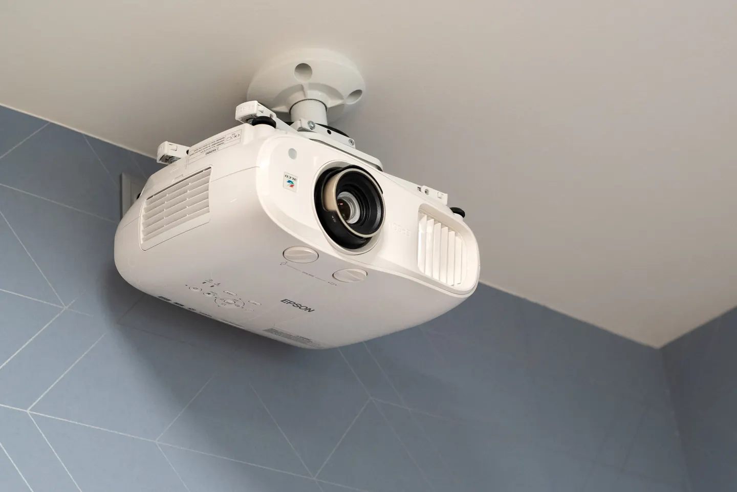 Mennyezetre konzollal rögzített fehér projektor, látható optikával és csatlakozásokkal; kék fal a háttérben.