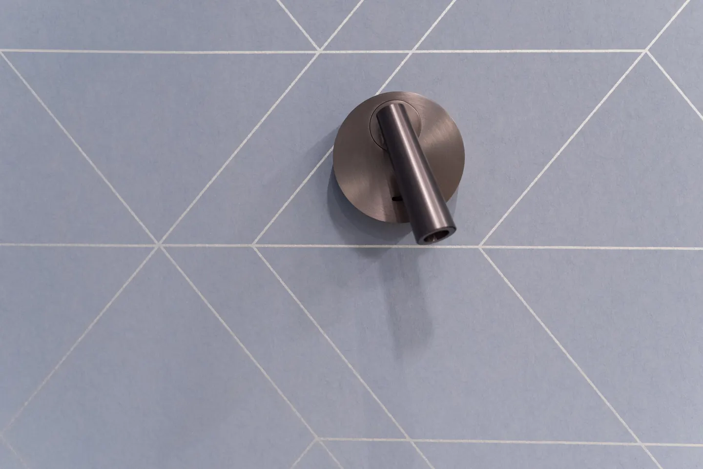 Sötét fémes, hengeres fali olvasólámpa kék geometrikus mintájú falon.